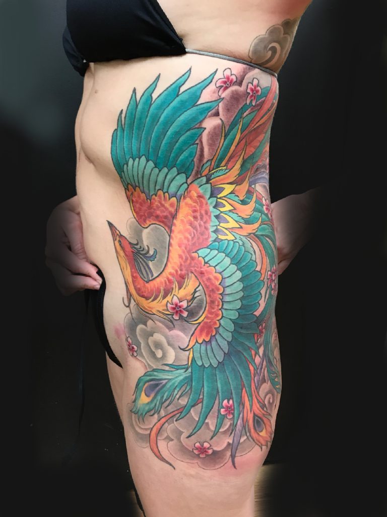 A vibrant, multicolored phoenix tattoo covering a significant portion of a person's side torso. The design includes flowers and clouds against a black background.