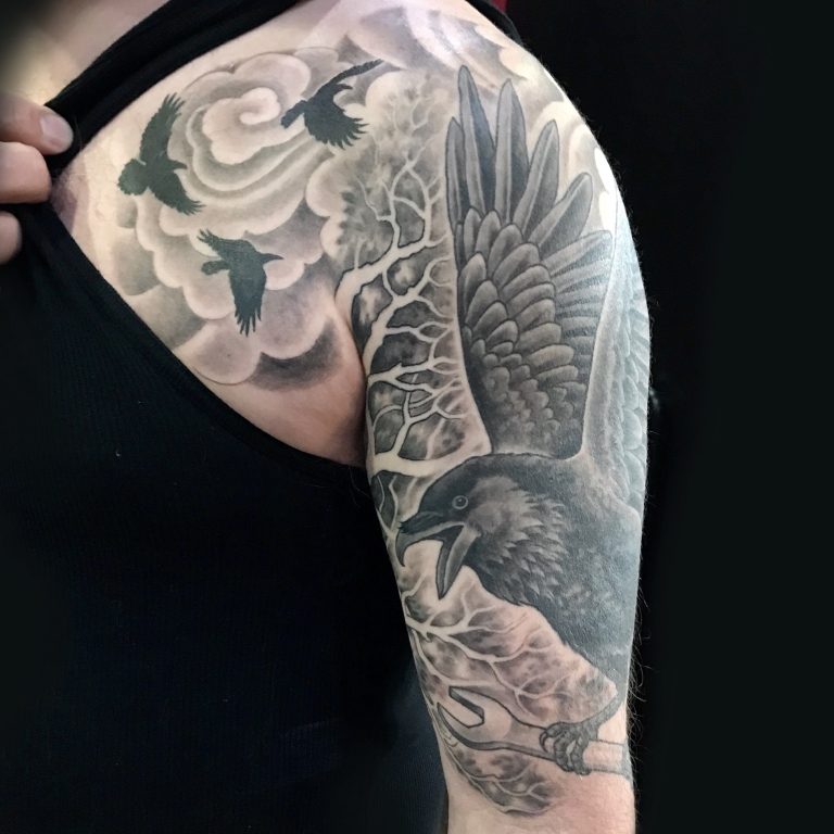 A person displays a shoulder tattoo of a raven with outstretched wings, surrounded by clouds and flying birds.