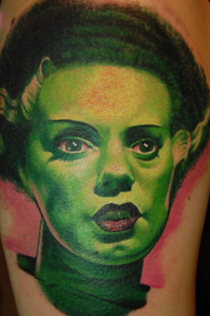 Tattoo of a woman's face with green skin, dark hair with white streaks, and red lips, on a person's skin with a pink background.