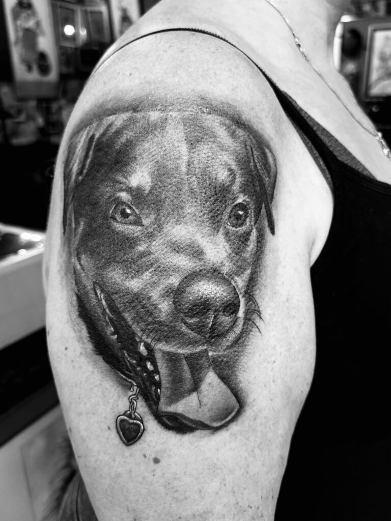 A realistic black and gray tattoo of a dog's face with its tongue out is inked on a person's upper arm.