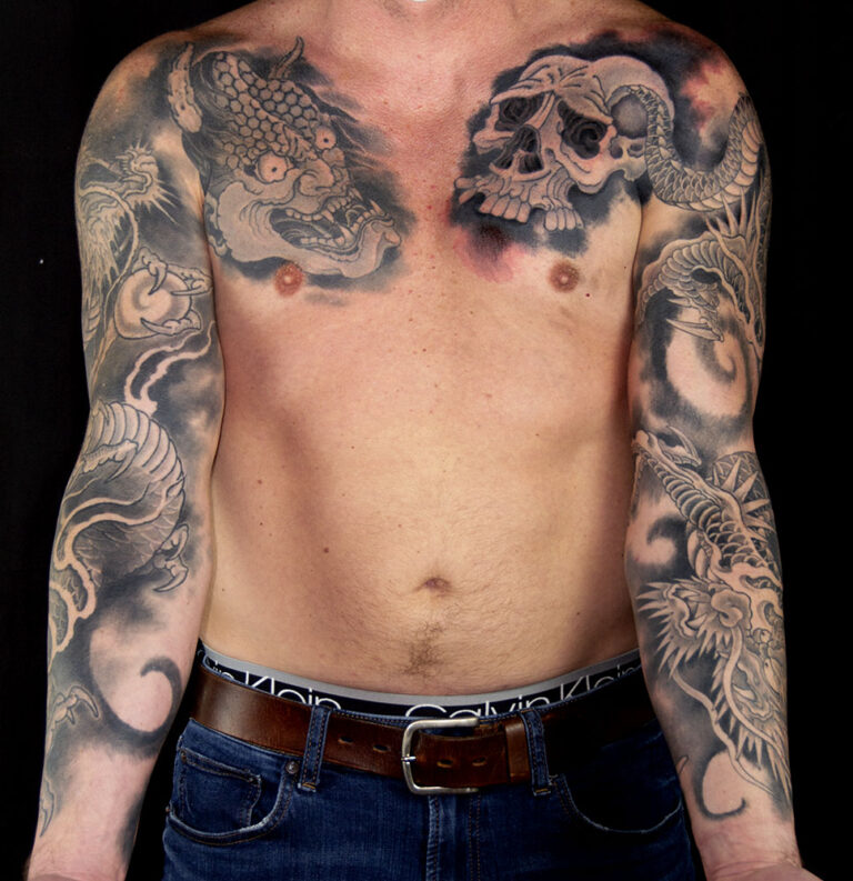 A shirtless person with detailed black and grey tattoos of a dragon and a skull on their chest and full sleeve tattoos on both arms.