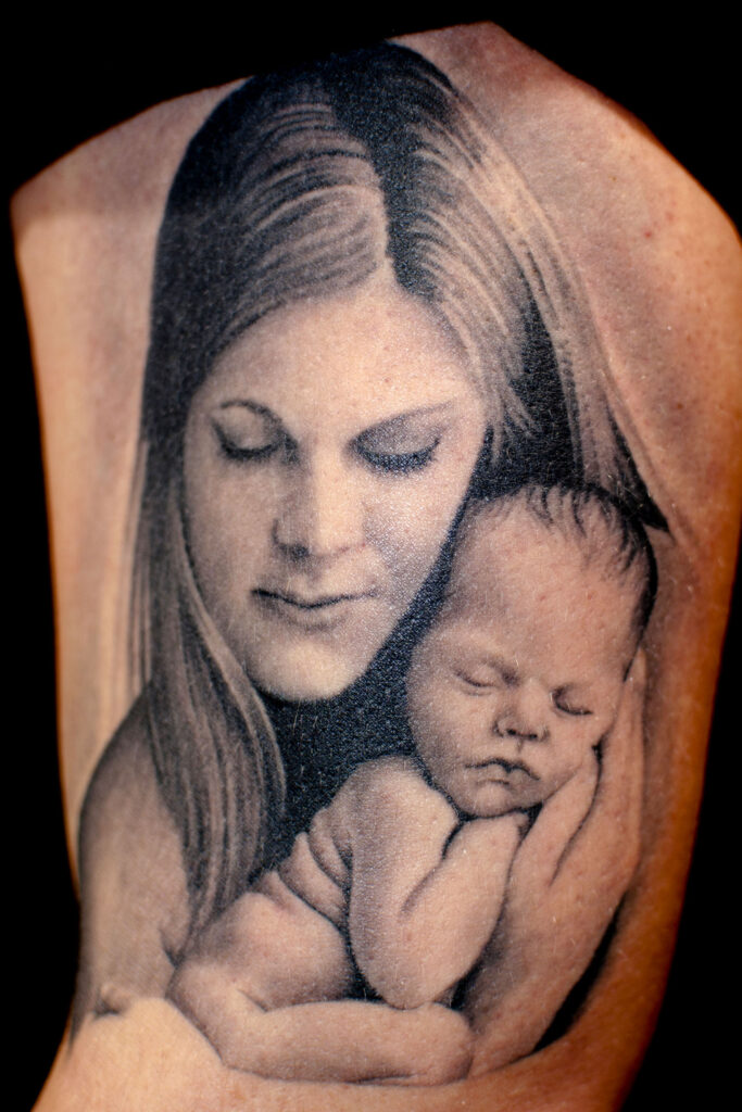 Black and gray tattoo showing a woman gently cradling a sleeping baby in her arms, both with calm expressions.