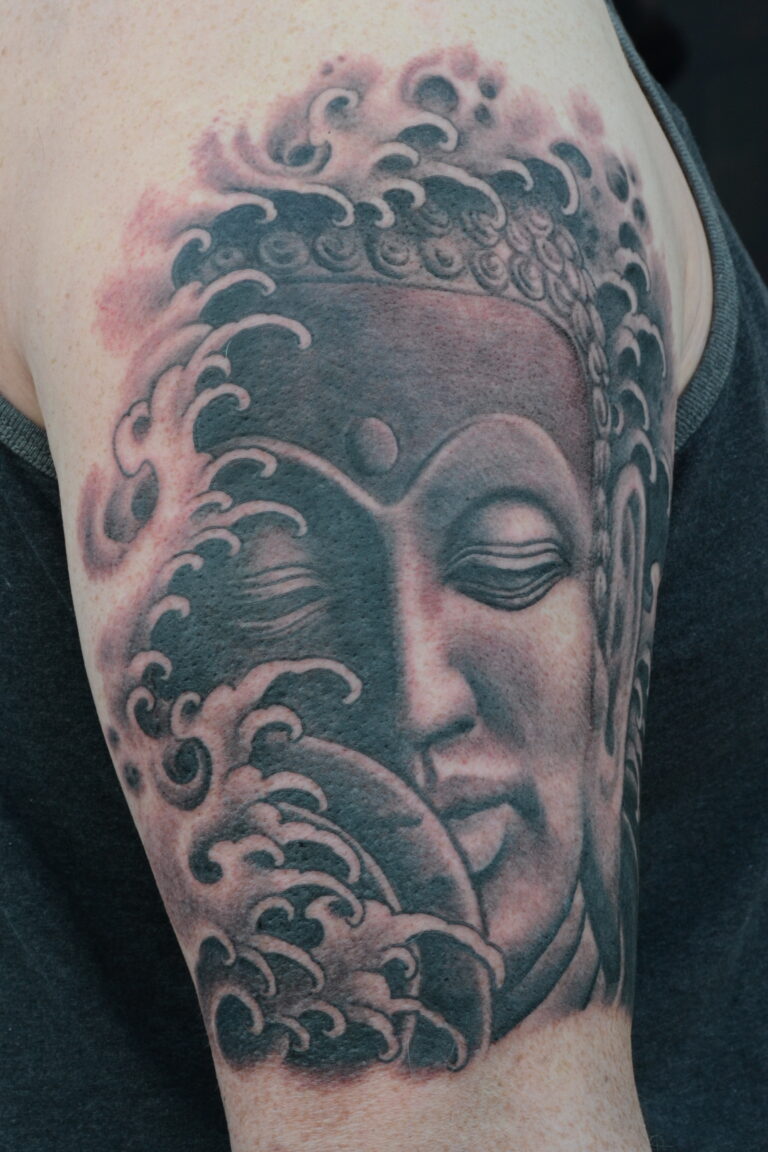 Tattoo of a serene Buddha face surrounded by stylized ocean waves on an upper arm.