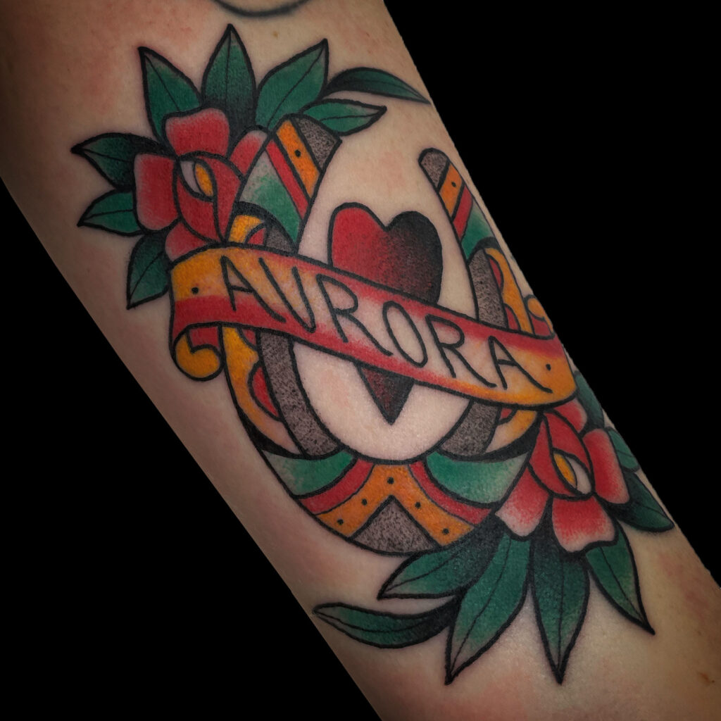 Colorful tattoo featuring a heart, horseshoe, and banner with the name "Aurora," surrounded by flowers and leaves.
